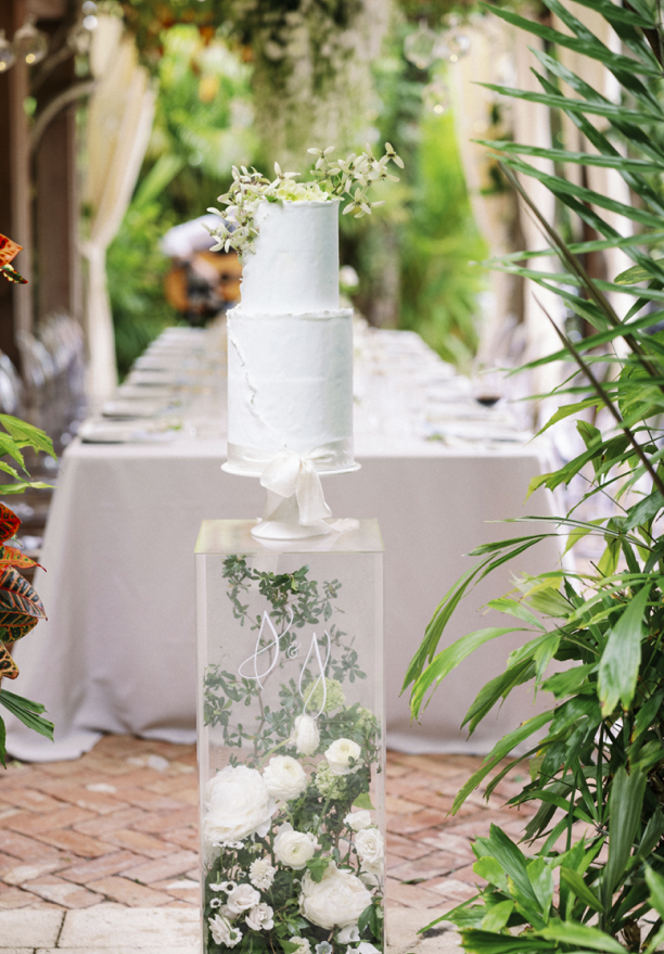 Beautiful white wedding cake covered in flowers sits on a pillar in front of a long table at The Escalante Hotel