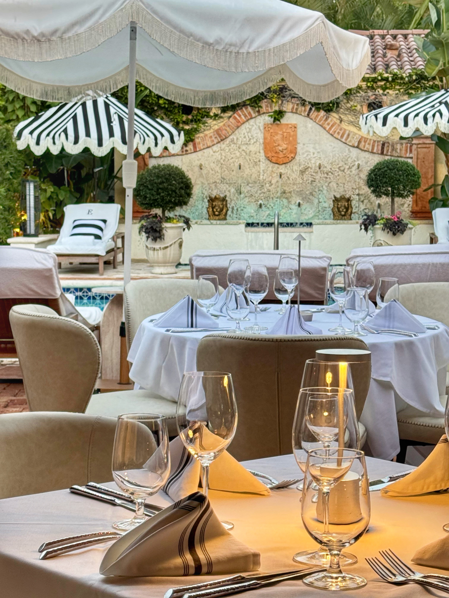 A fine dining setting with white tablecloths, wine glasses, and outdoor seating under umbrellas, overlooking a pool.